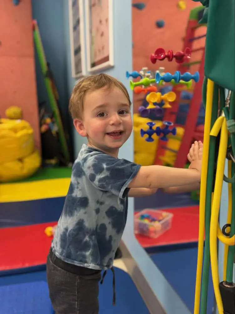 Smiling toddler standing and holding onto a climbing wall in the therapy gym